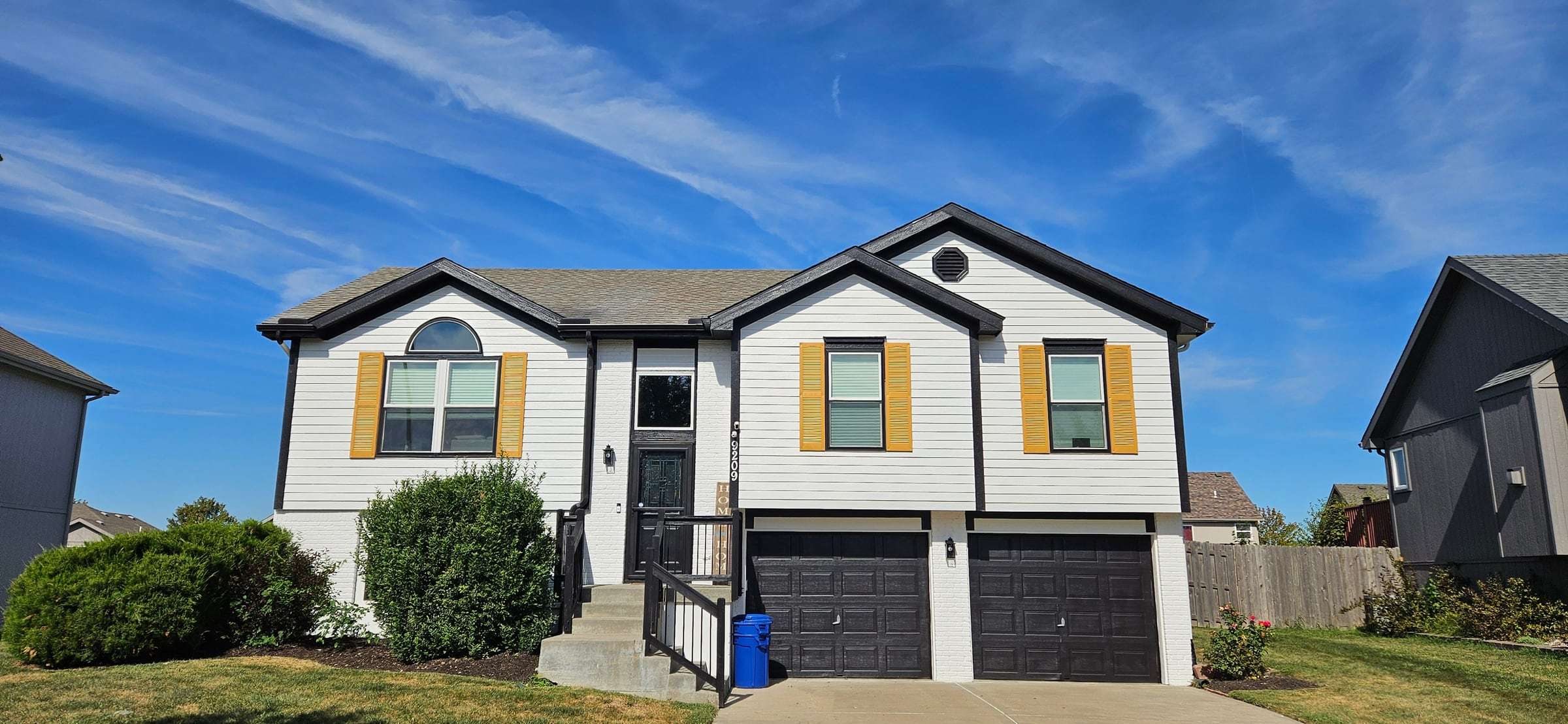 Split-level house after repainting: white siding, black trim and black garage doors, gold shutters
