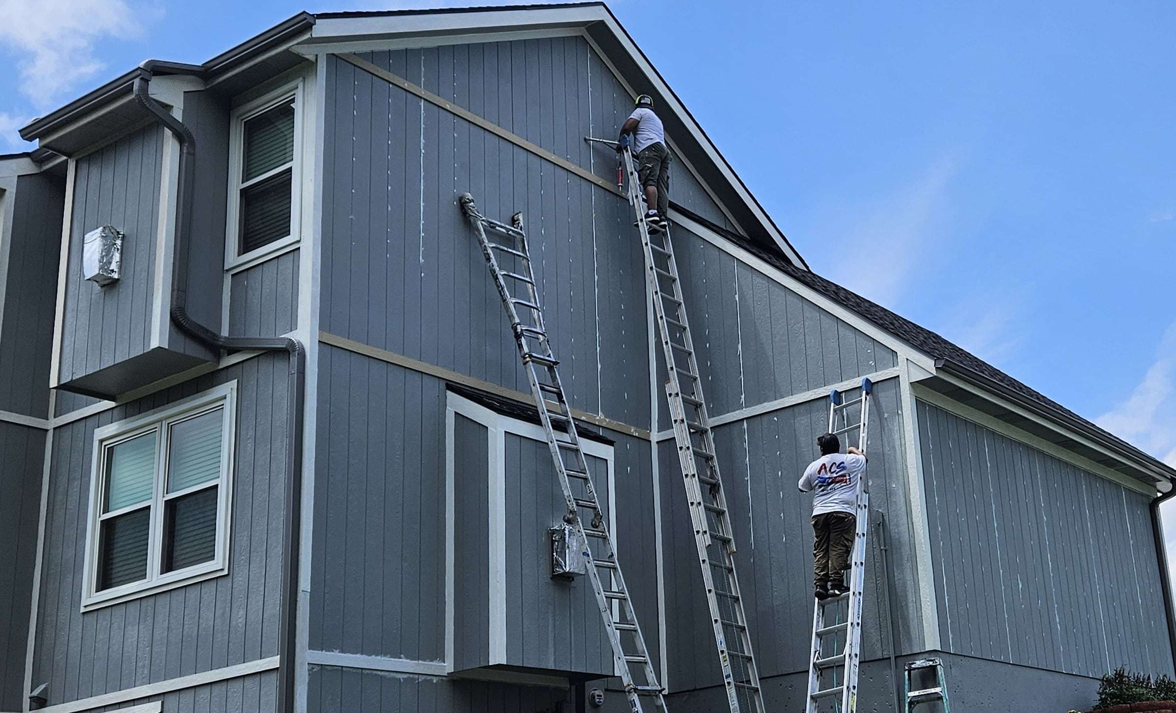 Two crew members on extension ladders caulking seams on gray-blue panel siding