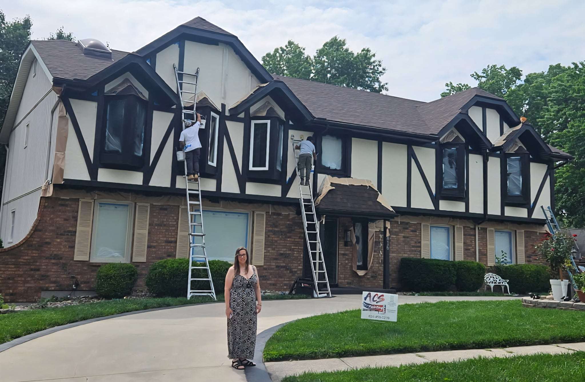 Ashlie Santini in the driveway of a Tudor-style house while two painters work on ladders behind her